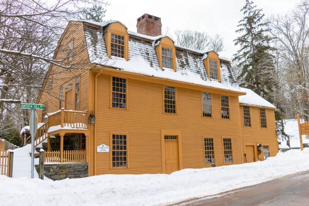 Two-story yellow colonial house with a shingled roof and dormer windows, surrounded by snow in winter.