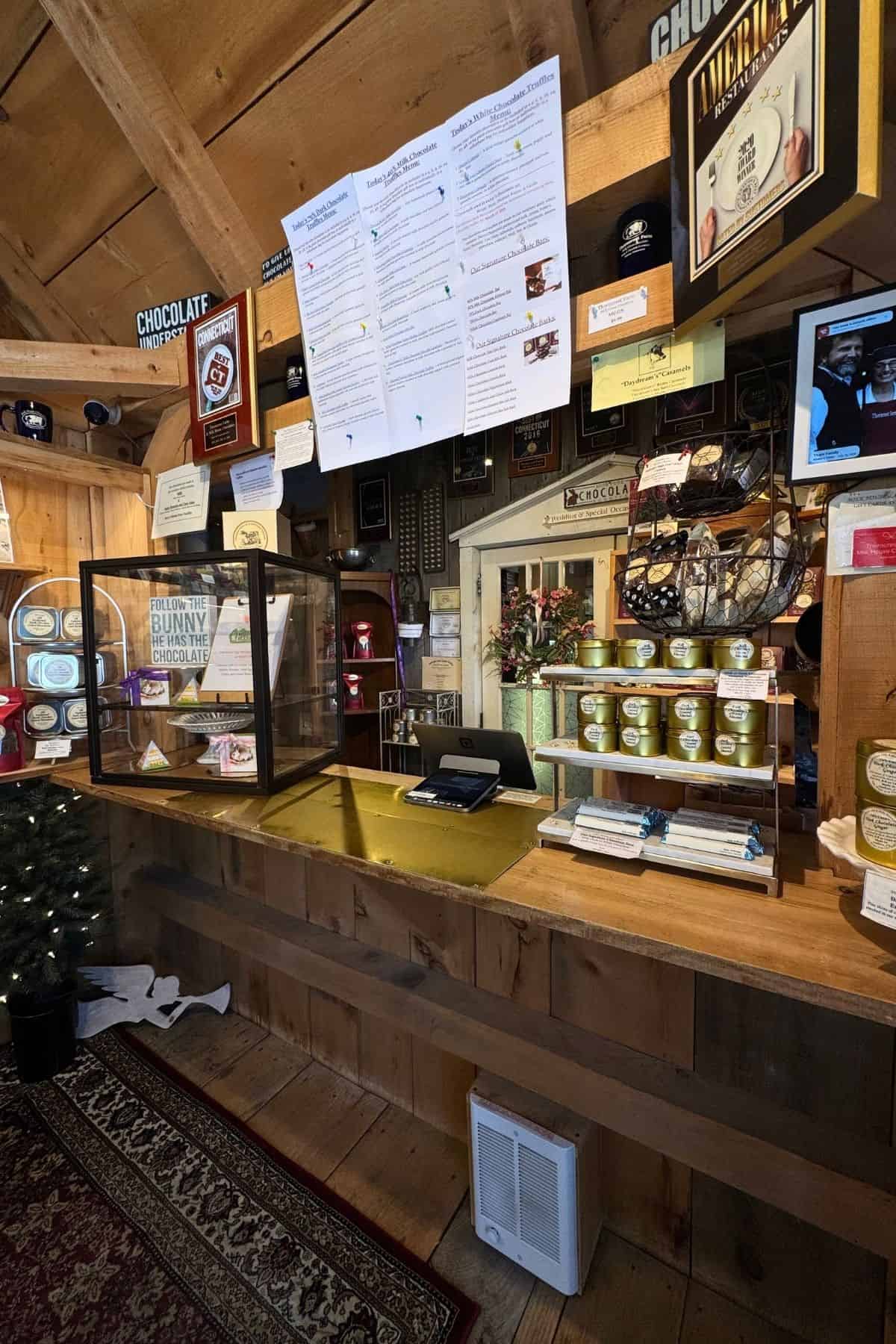 The interior of a rustic chocolate shop featuring a wooden counter, a glass display case, shelves filled with packaged sweets, and a menu posted on the wall.