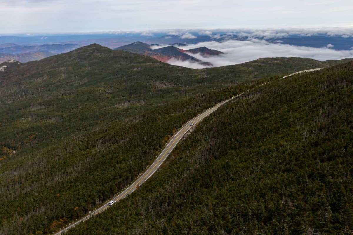 A winding road cuts through a forested mountain landscape with clouds settling in the valleys below.