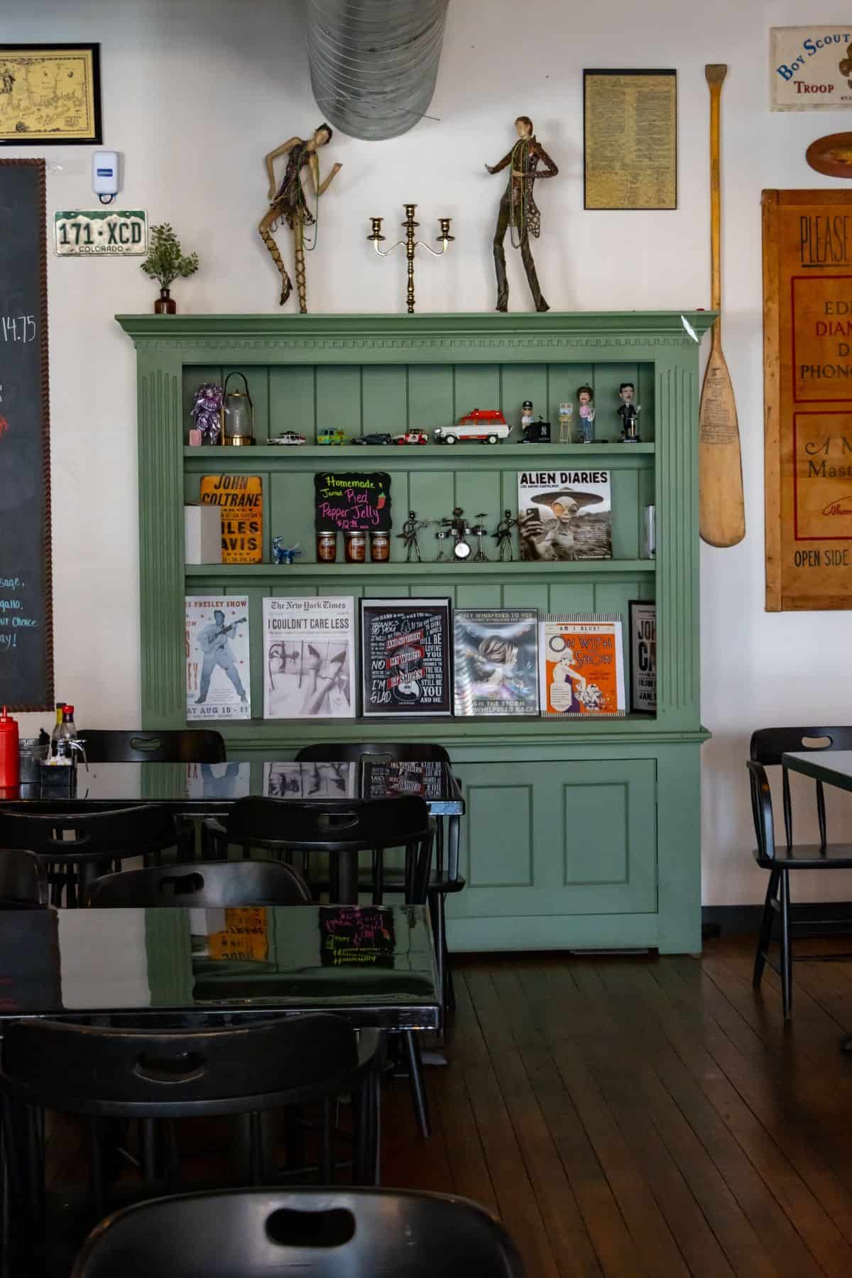 A large, green wooden hutch filled with books, small trinkets, and framed prints, positioned behind restaurant tables and chairs.