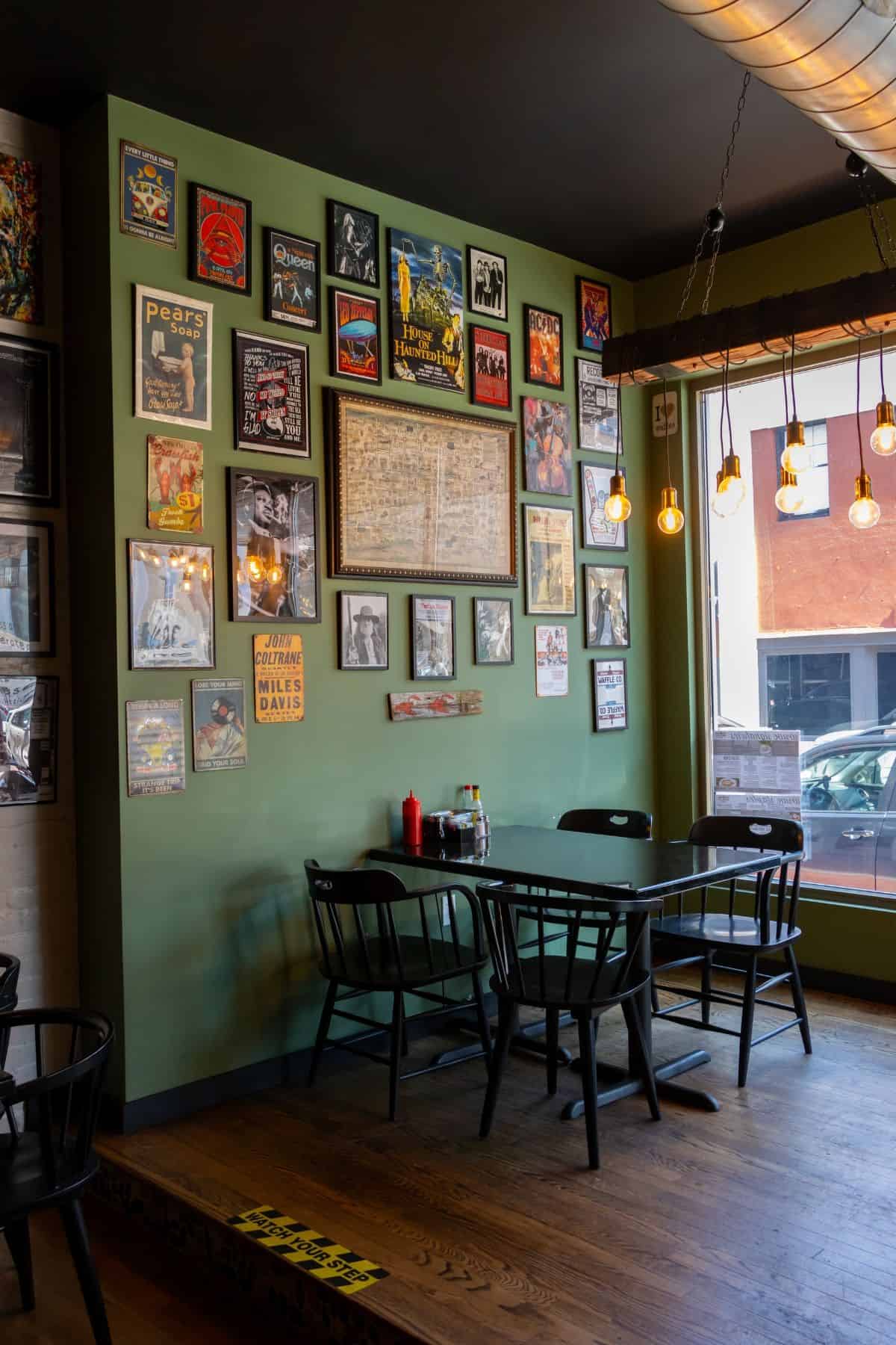 A small dining table with four black chairs against a sage green wall covered in vintage posters and a central framed map.