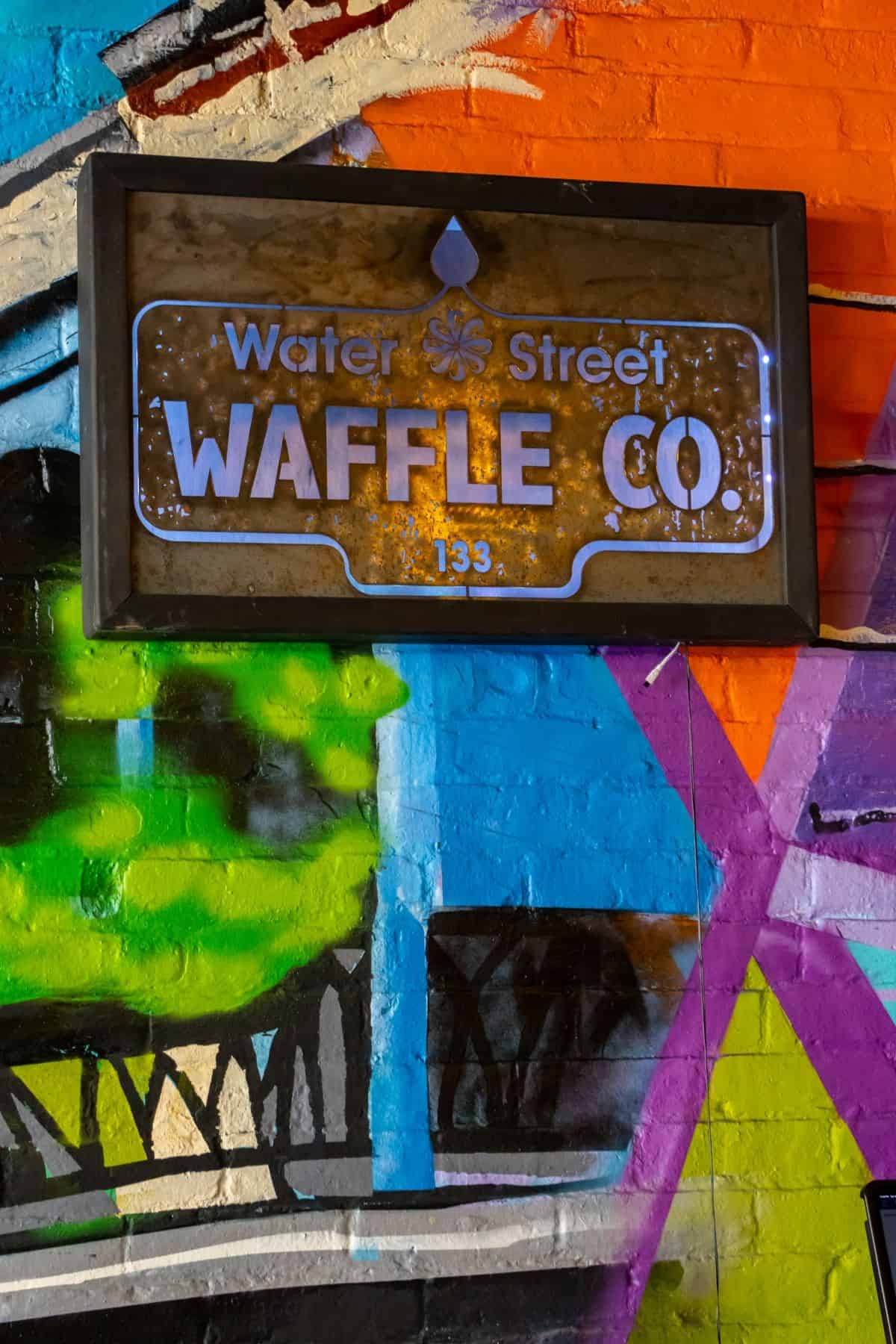 A backlit sign reading “Water Street WAFFLE CO. 133” mounted on a brick wall covered in colorful graffiti.