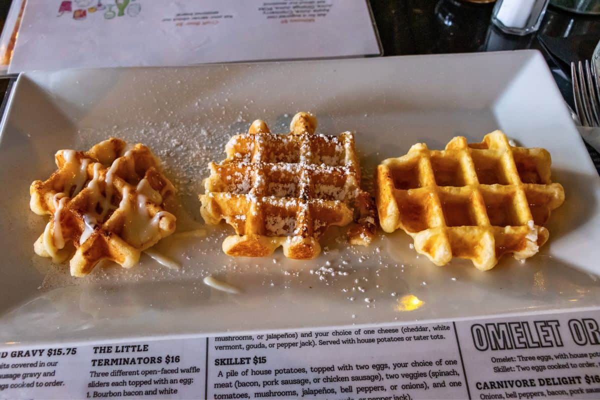 Three small waffles on a rectangular plate, drizzled with glaze, dusted with powdered sugar, and topped with syrup.