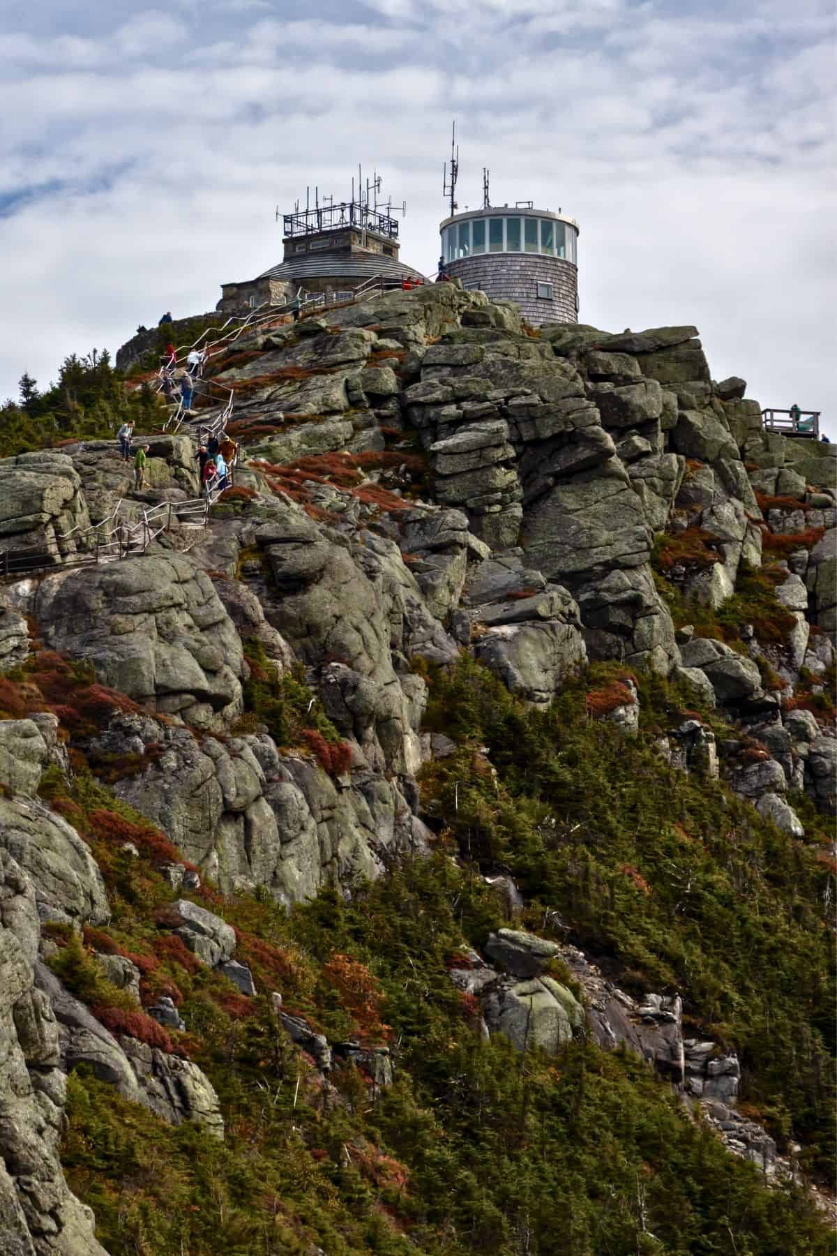 Hikers climb a wooden staircase up a steep, rocky mountain slope toward a stone observatory building.