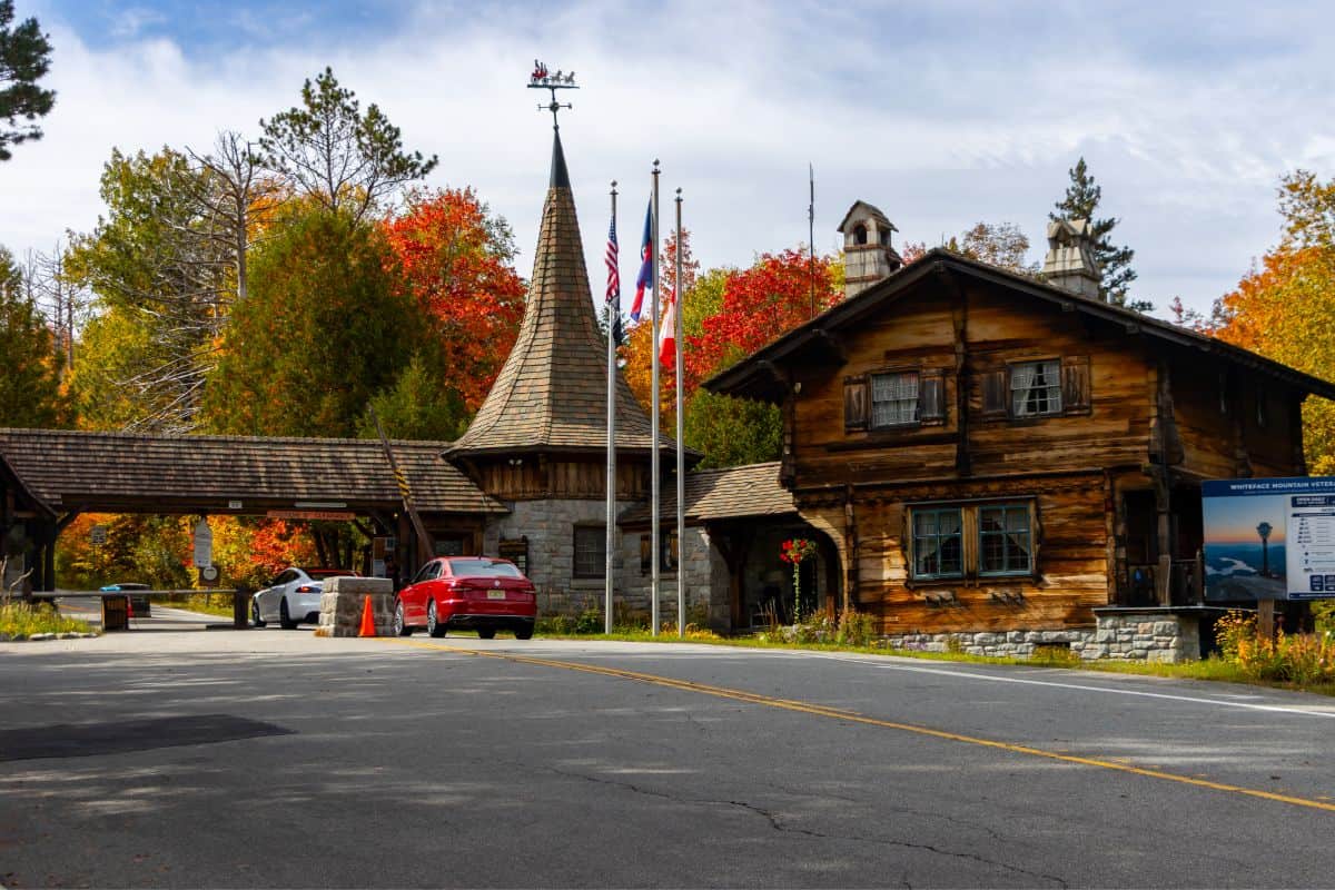 A gated entrance with a stone turret and a log building, surrounded by autumn trees, with cars passing through the gate.